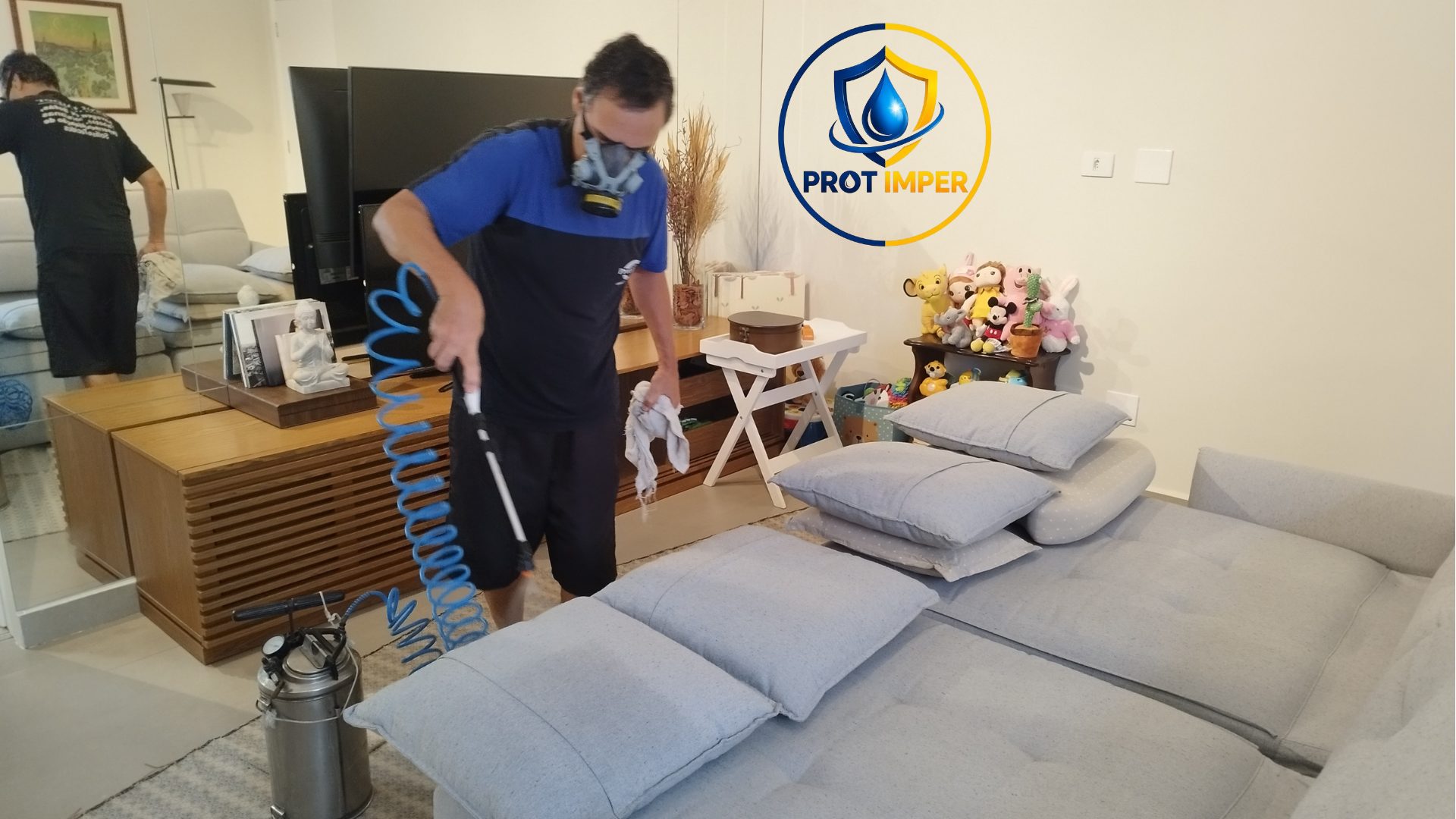 Técnico da PROT IMPER, de máscara, aplicando o impermeabilizante com pulverizador em um sofá cinza, com a marca PROT IMPER